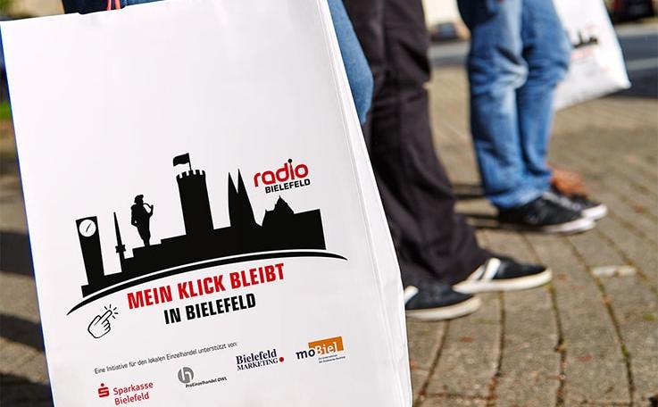 Papiertüte mit Aufdruck "Mein Klick bleibt in Bielefeld", dahinter Füße im Hintergrund