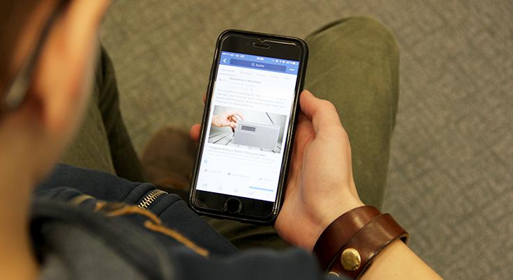 Blick über die Schulter eines jungen Mannes, der sich auf seinem Handy einen Facebook-Post ansieht.