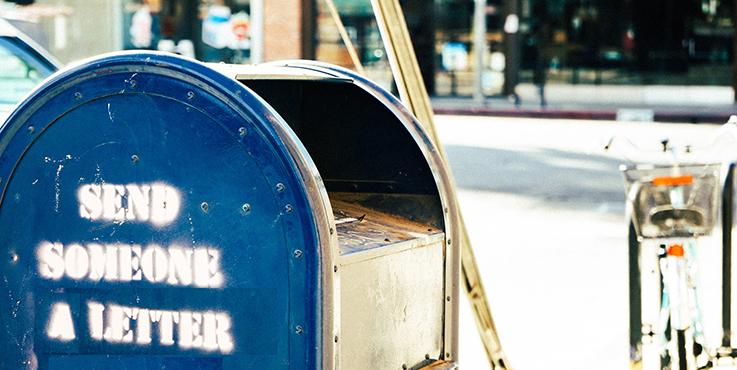 Nahaufnahme eines alten, blauen, amerikanischen Briefkastens, auf dessen Seite jemand den Schriftzug "Send someone a Letter" gesprüht hat.
