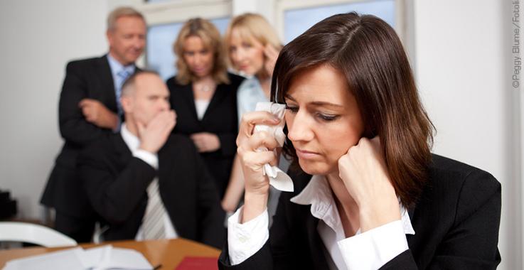 Frau im Büro trocknet Tränen, im Hintergrund lästert eine Gruppe Kollegen